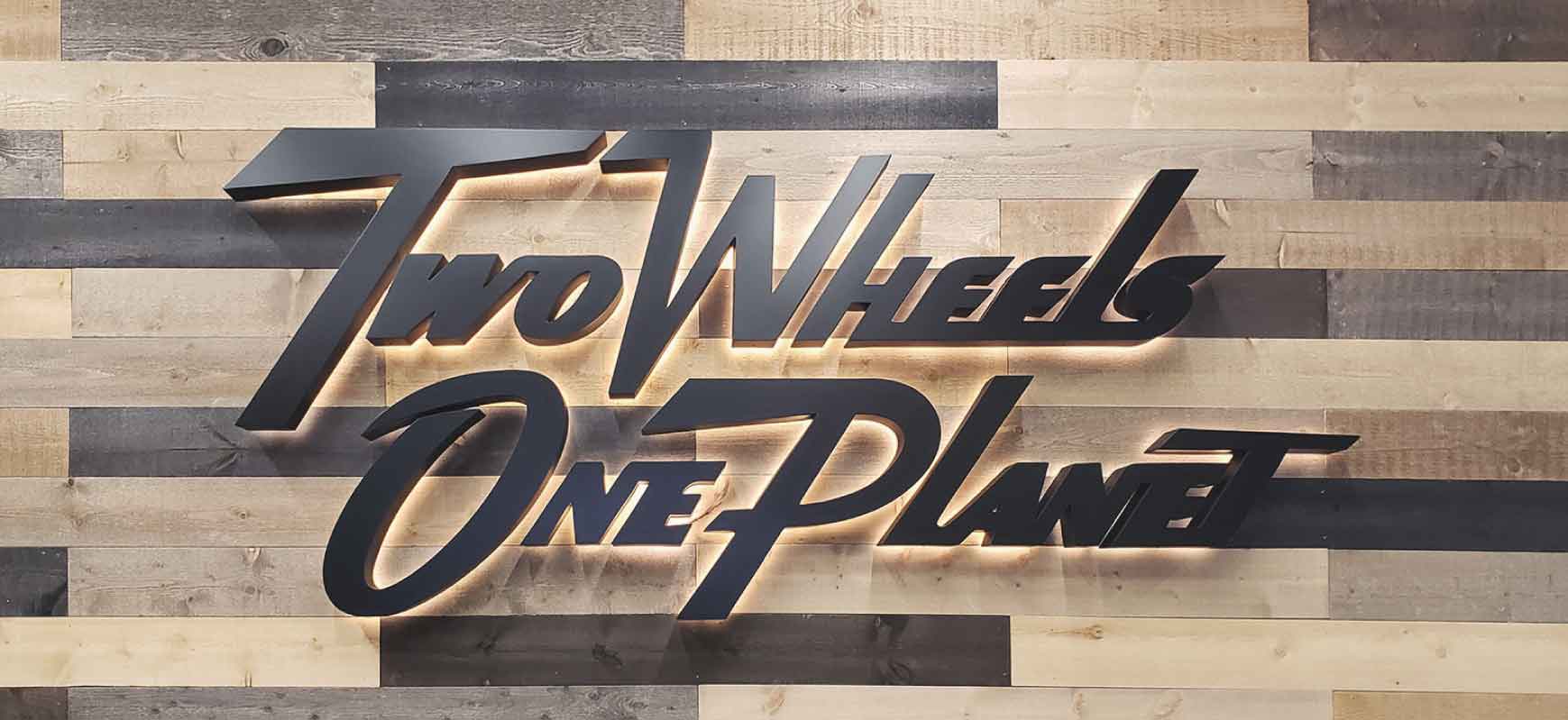 Two Wheels One Planet wall signage with black letters in italics made of lexan and aluminum